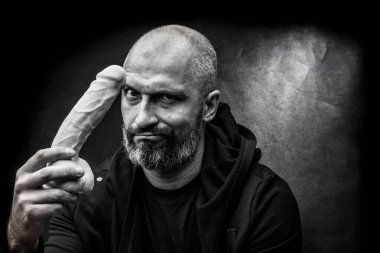 Kirli gri arka planda büyük gerçekçi bir dildosu olan kel sakallı bir adamın siyah beyaz portresi.