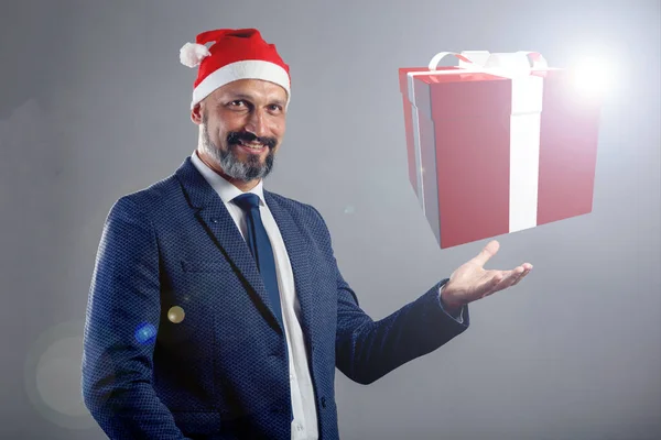 A bearded man in a blue jacket and a Christmas hat on a gray background holding a drawn 3D gift. Gift concept for new year, christmas.