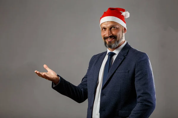 Bearded man in a blue jacket and a Christmas hat on a gray background holding an empty hand for editing with copy space