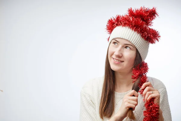 Funny young woman in white winter hat and sweater with red Christmas tinsel on a white background — Stock Photo, Image