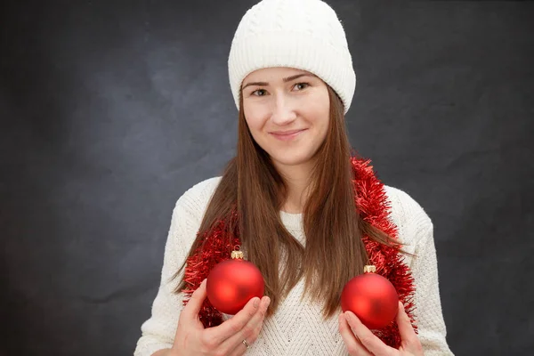 Lustige junge Frau mit weißem Winterhut mit rotem Weihnachtslametta und zwei Kugeln auf grauem Hintergrund — Stockfoto