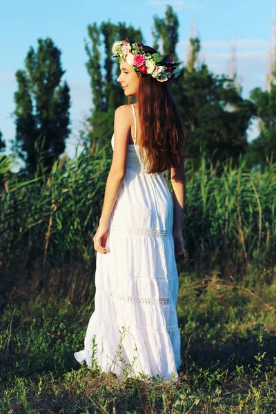 Femeie în rochie albă în picioare în câmp purtând coroană de flori . — Fotografie, imagine de stoc