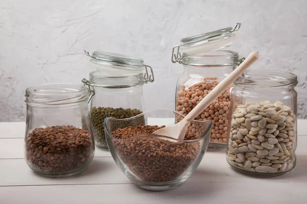 Raw lentils, white beans, mung bean and peas in a glass jar and buckwheat in a glass bowl and with a wooden spoon stand on a white wooden background