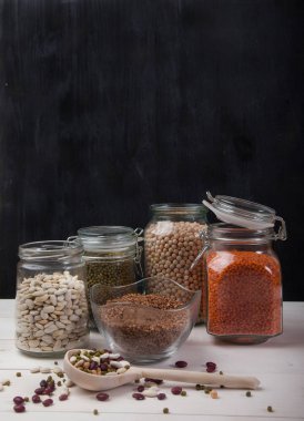 Kırmızı ve beyaz fasulye, bezelye, bezelye, kırmızı mercimek cam kavanozlarda ve siyah arka planda beyaz ahşap bir masada tahta kaşık..