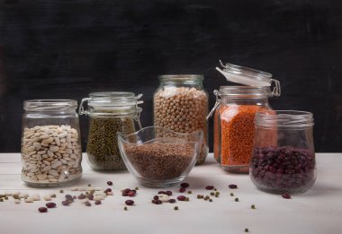 Kırmızı ve beyaz fasulye, bezelye, bezelye, cam kavanozlardaki kırmızı mercimek siyah arka plandaki beyaz ahşap bir masada duruyor.