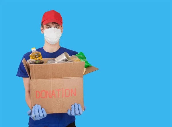 Young man, a volunteer in a medical mask and rubber gloves holds a cardboard box with humanitarian aid products on a blue background. Social assistance