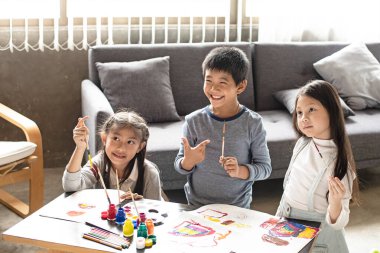 İki kız kardeş ve genç çocuk su rengini kağıda boyuyor, birlikte aktiviteler yapıyorlar, oturma odasında, öğrenme zamanı.