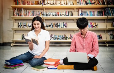 Genç öğrenci kitap okuyor, arkadaşı dizüstü bilgisayar kullanıyor, kütüphanede aktivite yapıyor. Bulanık ışık altında.