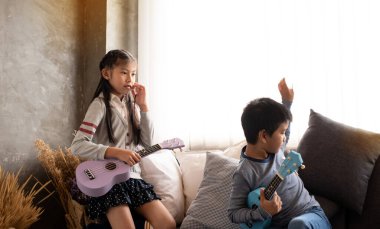 İki çocuk kanepede oturuyor, birlikte ukulele çalmaya hazırlanıyorlar, stüdyoda, bulanık ışık etrafında.