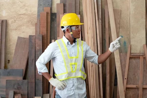 Handsome man wearing protective glasses and yellow helmet,point finger to left side,standing in front of wood pile,at factory