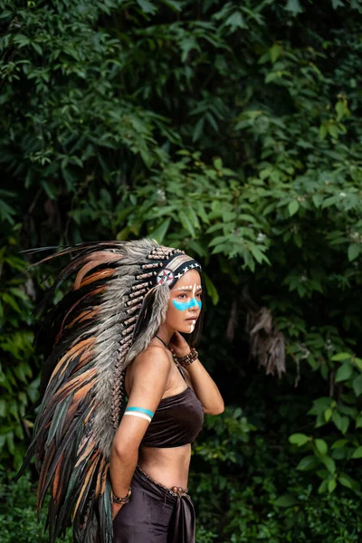 这个漂亮的女人头戴鸟类头饰羽毛 脸上涂着蓝色 在森林里摆姿势的模特画像 周围光线模糊 — 图库照片