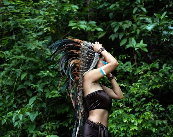 The beautiful woman raising hand touch headdress made from feathers of birds. painted blue color on her cheek,portrait of model posing,Jungle in forest,blurry light around