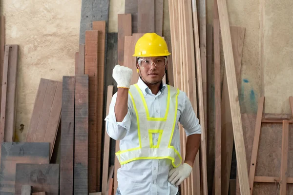 Handsome man wearing protective glasses and yellow helmet,raised right fist up with determind eyes,standing in front of wood pile,at factory