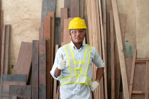 Handsome man wearing protective glasses and yellow helmet,raise thumpnail up,standing in front of wood pile,at factory