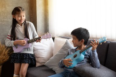 Küçük çocuğun ukulele çaldığı ve kanepenin solundaki bulanık tatlı kıza gülümsediği seçici odak noktasında, ev stüdyosunda, bulanık ışık etrafında.
