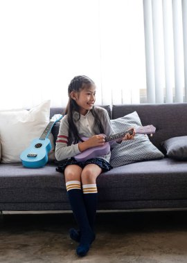 Kanepede oturan küçük kız, mutlu bir şekilde ukulele çalıyor, bulanık bir ışık saçıyor.