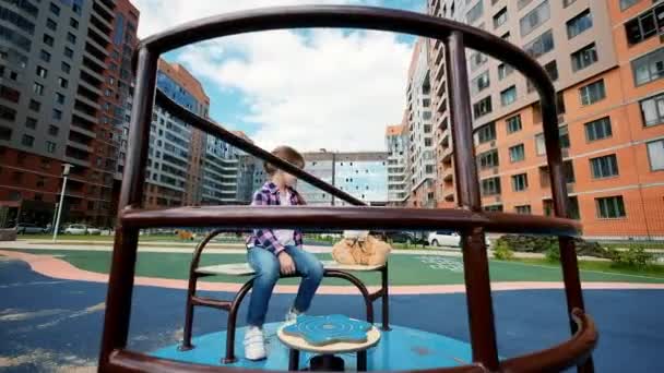 Little girl play on park playground with a carousel. A lonely child is riding a carousel with toy in yard on summer. Pretty child spins on a carousel. Adorable girl has fun at playground. — стоковое видео