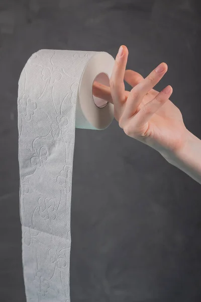 On the finger of a woman's hand holding a roll of toilet paper on a gray background, close-up