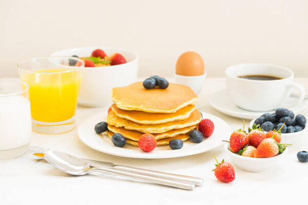 Здоровый завтрак с блинами и гранолой с черникой и клубничным и черным кофе, молоком и апельсиновым соком на белом фоне каменного стола
