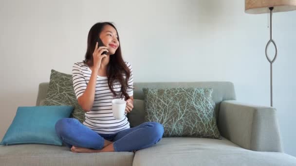 美しいアジアの女性が熱いコーヒーを飲みながら家でひとりで過ごし電話で話している映像 — ストック動画