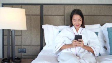 Yatakta akıllı telefon kullanan bornozlu güzel Asyalı kadının görüntüleri.