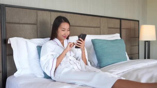 穿着浴衣的美丽的亚洲女人在床上用智能手机的镜头 — 图库视频影像
