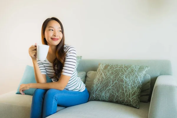 소파에 커피 잔을 들고 있는 아름다운 젊은 아시아 여성의 사진 — 스톡 사진