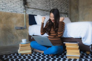 Genç Asyalı kadın elinde kahve fincanı ve kitapla dizüstü bilgisayar kullanıyor.