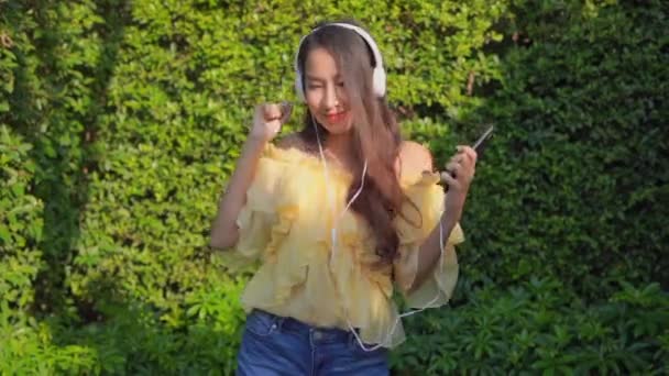 庭でヘッドフォンで音楽を聴いている美しいアジアの女性の映像 — ストック動画