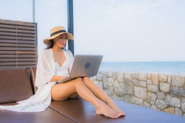 Plaj denizi çevresinde çalışmak için dizüstü bilgisayarı kullanan Asyalı genç bir kadının portresi.
