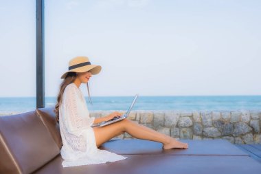 Plaj denizi çevresinde çalışmak için dizüstü bilgisayarı kullanan Asyalı genç bir kadının portresi.