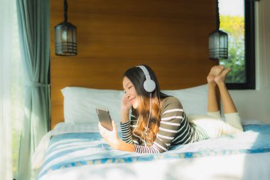 Asyalı genç bir kadının portresi yatak odasının içinde müzik dinlemek için kulaklıklı akıllı cep telefonu kullanıyor.