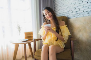 Genç Asyalı kadının portresi kanepeye oturur ve oturma odasının içinde kahve fincanı ile kitap okur.