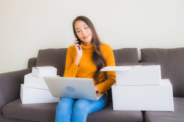 Asyalı genç bir kadının portresi, oturma odasındaki koltukta paket kutusuyla çalışmak için dizüstü bilgisayar kullanıyor.