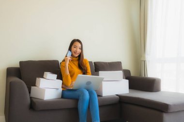 Asyalı genç bir kadının portresi, oturma odasındaki koltukta paket kutusuyla çalışmak için dizüstü bilgisayar kullanıyor.
