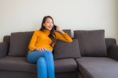 Genç Asyalı kadının oturma odasında yastıklı akıllı cep telefonu kullandığı portre.
