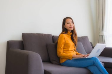 Genç Asyalı bir kadının oturma odasında çalışmak için dizüstü bilgisayar defteri kullanmasının portresi.