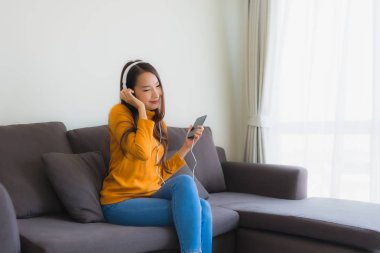 Genç Asyalı kadın oturma odasındaki kanepede müzik dinlemek için akıllı cep telefonu ve kulaklık kullanıyor.