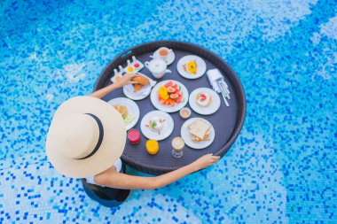 Asyalı güzel bir kadının portresi yüzme havuzunda yüzen kahvaltı tepsisiyle mutlu bir gülümseme