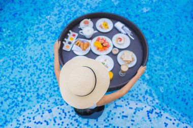 Asyalı güzel bir kadının portresi yüzme havuzunda yüzen kahvaltı tepsisiyle mutlu bir gülümseme