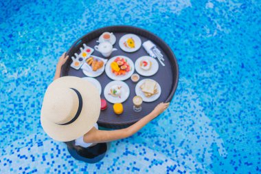 Asyalı güzel bir kadının portresi yüzme havuzunda yüzen kahvaltı tepsisiyle mutlu bir gülümseme