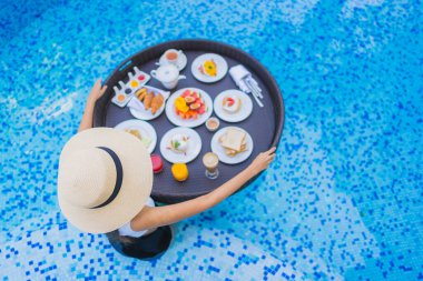 Asyalı güzel bir kadının portresi yüzme havuzunda yüzen kahvaltı tepsisiyle mutlu bir gülümseme