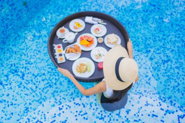 Asyalı güzel bir kadının portresi yüzme havuzunda yüzen kahvaltı tepsisiyle mutlu bir gülümseme