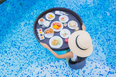 Asyalı güzel bir kadının portresi yüzme havuzunda yüzen kahvaltı tepsisiyle mutlu bir gülümseme