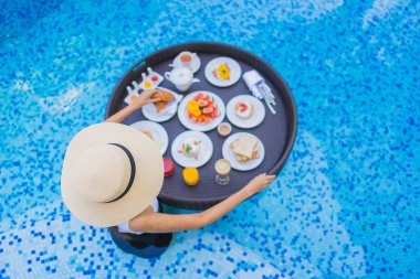 Asyalı güzel bir kadının portresi yüzme havuzunda yüzen kahvaltı tepsisiyle mutlu bir gülümseme