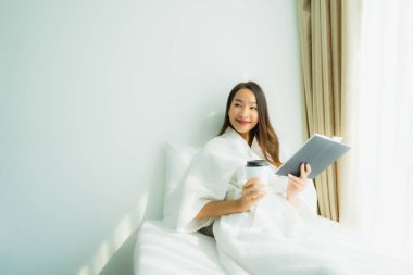 Genç Asyalı kadın, elinde kahve fincanıyla yatak odasında kitap okuyor.