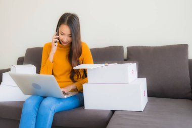 Asyalı genç bir kadının portresi, oturma odasındaki koltukta paket kutusuyla çalışmak için dizüstü bilgisayar kullanıyor.