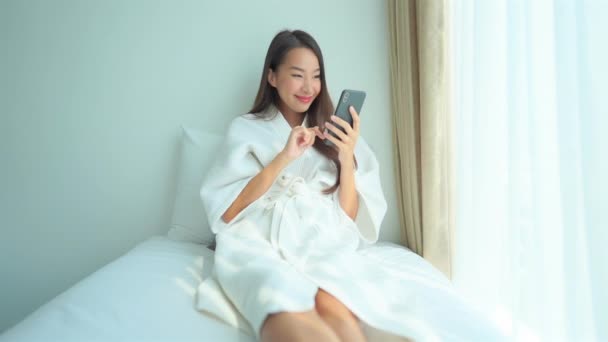 寝室のインテリアでベッドの上でスマートフォンを使用して肖像画の美しい若いアジアの女性 — ストック動画