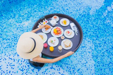 Asyalı güzel bir kadının portresi yüzme havuzunda yüzen kahvaltı tepsisiyle mutlu bir gülümseme