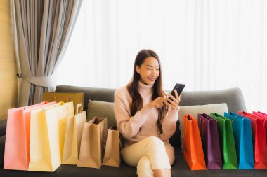 Portre güzel genç Asyalı kadın dizüstü bilgisayar ya da akıllı cep telefonu kullanarak oturma odasındaki alışveriş çantasının etrafındaki kanepede alışveriş yapıyor.